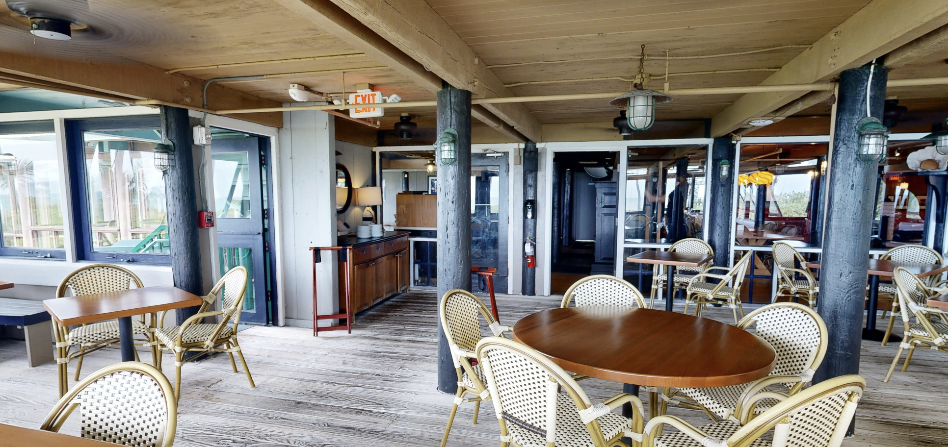 Laser-scan photo of the Sea Watch covered ground-floor deck dining area, showing timber pile columns, the wood-plank ceiling and floor, ocean-view windows, and bistro tables and chairs.
